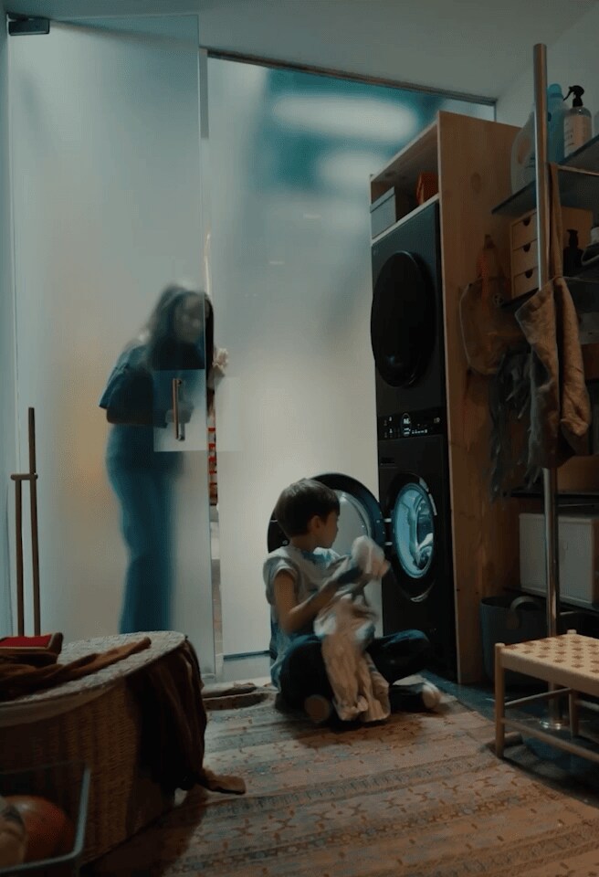 Một người mẹ và con trai cùng nhau sử dụng máy giặt LG AI, vặn núm xoay để kích hoạt chức năng AI Wash. Một người đàn ông đang sử dụng máy tính xách tay LG gram cũng xuất hiện trong cùng một phân cảnh.