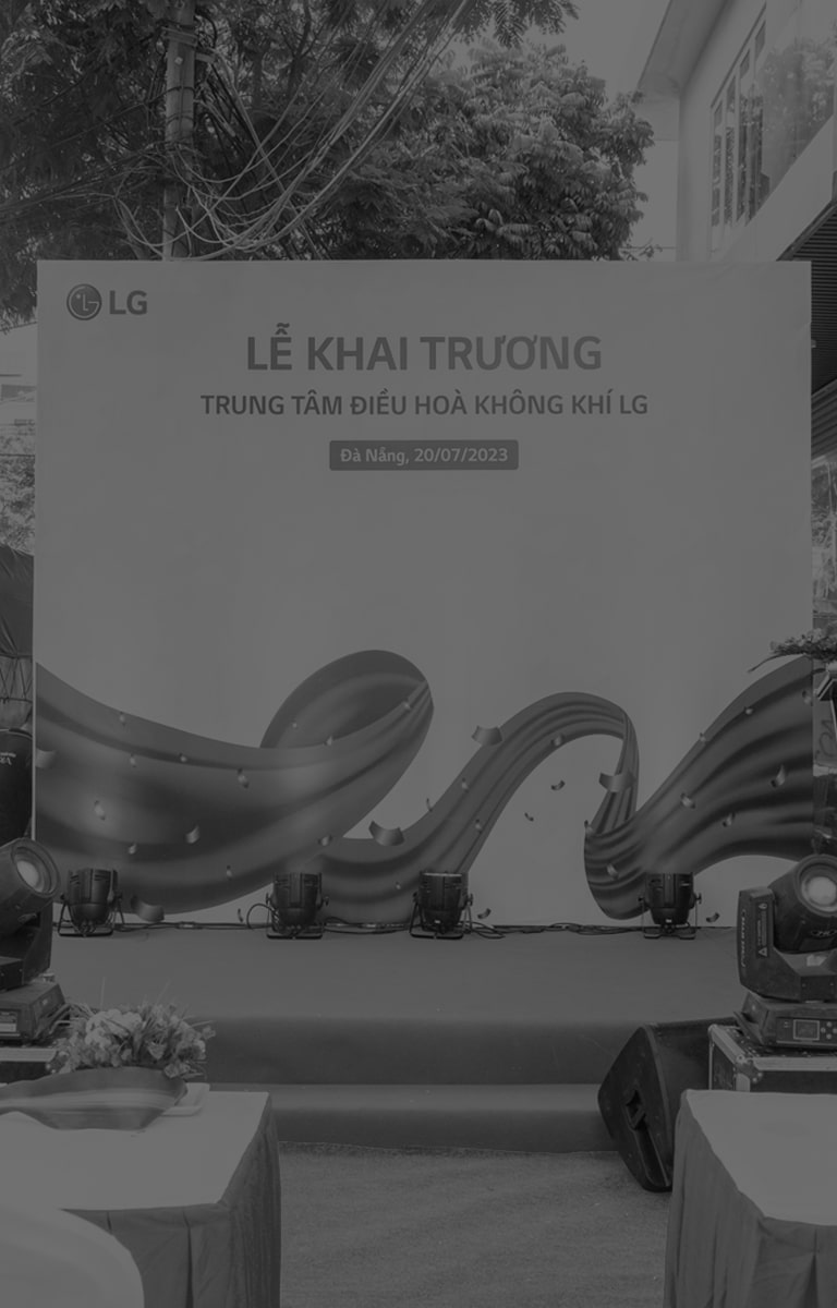 Dưới tấm biển lớn 'Học viện LG' ở góc trên bên trái, mọi người đang đứng thành nhiều hàng trên bục trong lễ khai mạc. Bên trái họ là những bó hoa lớn màu đỏ.