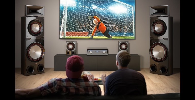 LG MR11 is placed in the living room. LG TV is hung on the wall, between two giant tall boy speakers, displaying a soccer game. Two people are watching the game.