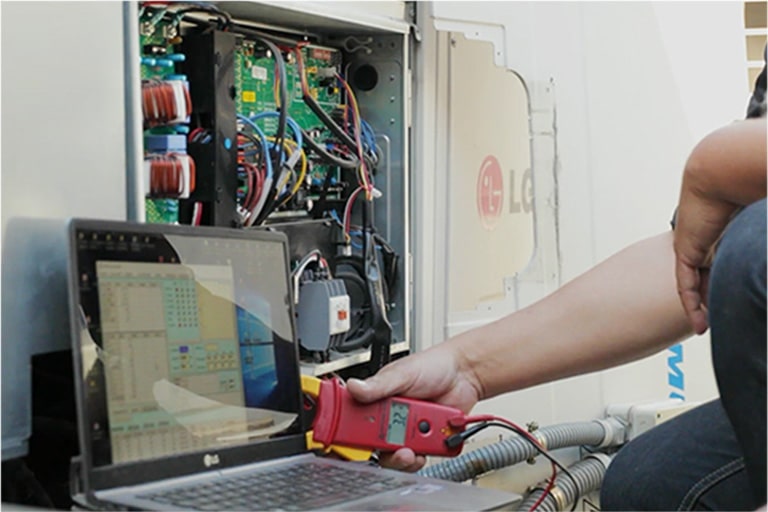 A person is using a red rectangular tool connected to a laptop, diagnosing an open LG outdoor unit, its internal wiring fully exposed.