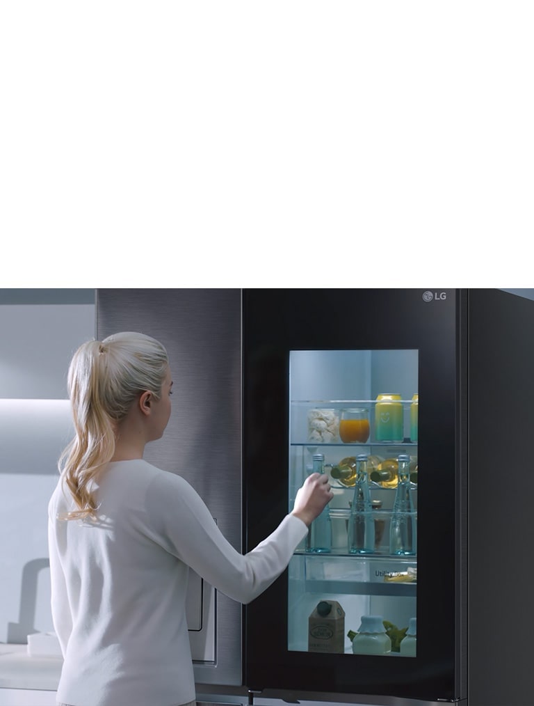 A video shows a woman approach her InstaView refrigerator and knock twice. The interior lights up and she can see the contents of her fridge without opening the door. The view zooms in to focus on the drinks in the door and then zooms out to see the woman from behind as she opens the door and grabs a drink.