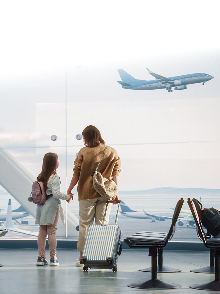 A woman and a child holding hands, standing at an airport gate with luggage, looking at airplanes outside.