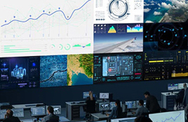 ontrol room with multiple large display screens showing data charts, maps, and monitoring dashboards while people work at computer stations.