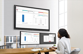 Woman working at a desk in an office, analyzing data displayed on multiple monitors including a large wall-mounted screen.