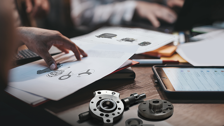 Hands pointing at documents and blueprints on a table, with a disassembled compressor part nearby. Engineers collaborating on product development.