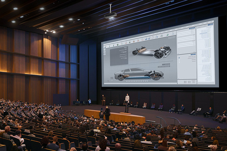 A large auditorium filled with people, focusing on a presenter standing in front of a massive screen displaying a detailed 3D car design. An overhead projector is mounted on the ceiling, projecting the visuals clearly across the spacious hall.