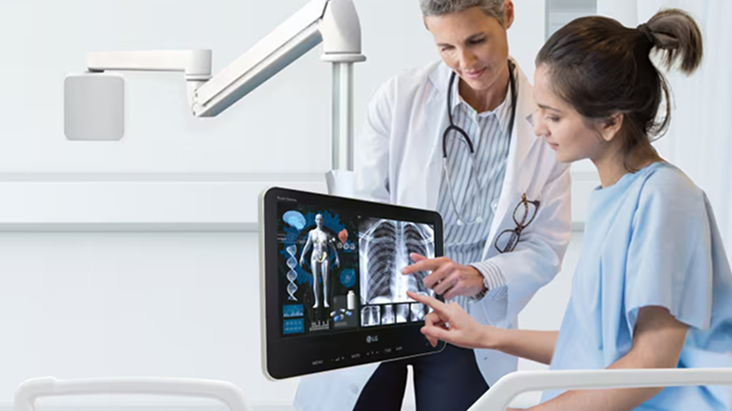 A doctor and a patient in a hospital room review medical scans displayed on a touchscreen monitor beside the patient's bed.