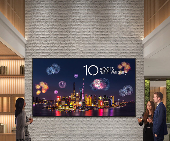 Large LG digital signage mounted on a stone wall in a modern lobby, displaying a city skyline with fireworks and the text '10 Years Anniversary,' while people stand nearby viewing the screen