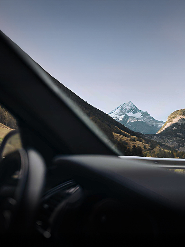 View of a snow-capped mountain and forested hills through a car window while driving along a guardrailed road.
