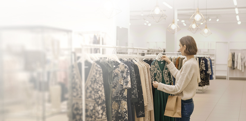 Woman shopping in a clothing store, holding a green embroidered dress while carrying a shopping bag.