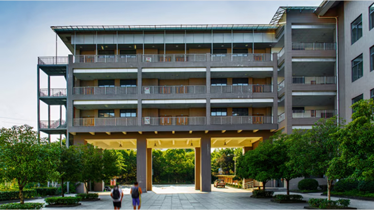 A modern apartment building with multiple balconies surrounded by trees and shrubs under a clear sky.