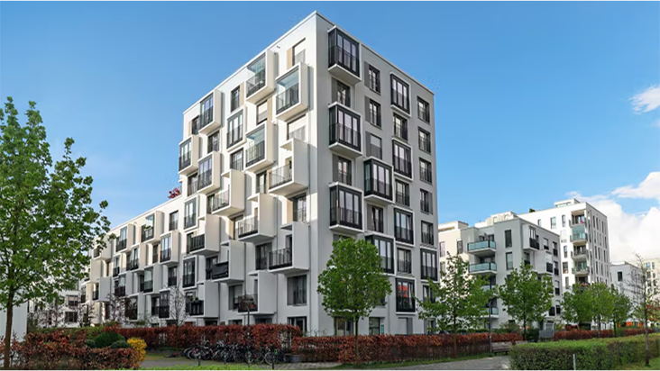 A modern apartment building with multiple balconies surrounded by trees and shrubs under a clear sky.