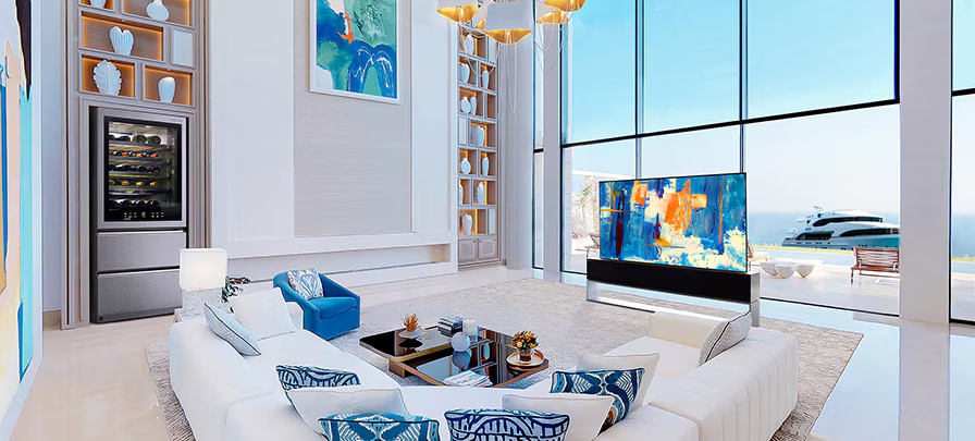 LG SIGNATURE Rollable OLED TV R and Wine Cellar in a white-and-blue-themed room in front of large windows with a view of the ocean.