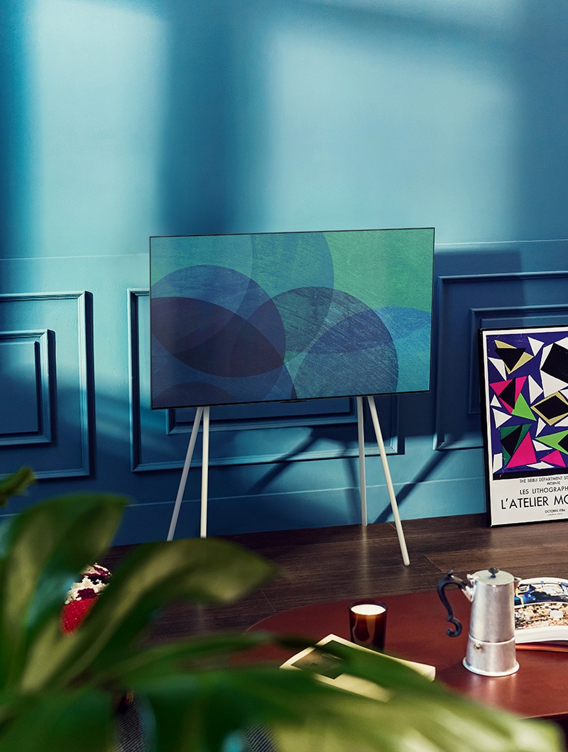 En una moderna habitación gris frente a una ventana que da a un bosque nevado hay un televisor LG OLED con soporte Gallery. En una colorida sala de estar con muebles de madera y tonos pastel hay un televisor LG OLED con soporte de suelo.