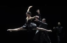 A female dancer is captured doing the splits mid-air, accompanied by a male dancer standing towards her. The two are clad in black costumes.