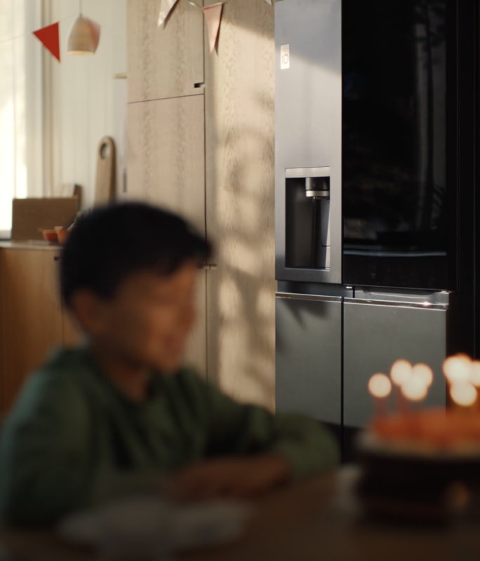 The child's face is faintly visible in front of the candlelit cake, and the LG InstaView refrigerator is clearly visible in the background. 